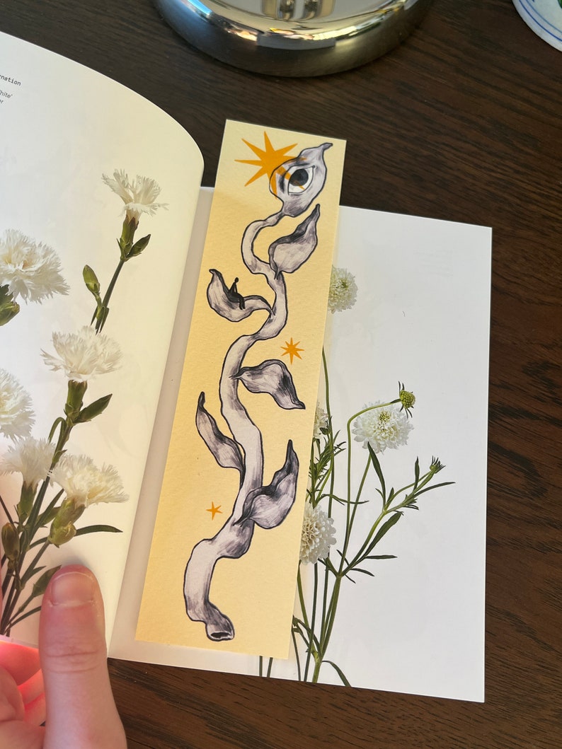 May include: A yellow bookmark with a black and white illustration of a vine with an eye and stars. The vine is stylized and has a whimsical feel.