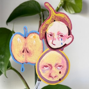 Puede incluir: Tres recortes de papel dibujados a mano de caras y una manzana están sujetos a una planta. El recorte superior es una cara con una cáscara de plátano en la cabeza. El recorte del medio es una manzana. El recorte inferior es una cara con un borde púrpura.