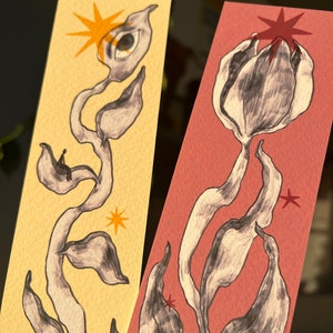 May include: Two hand-painted bookmarks with abstract floral designs. One bookmark is on a yellow background with a black and white flower with an eye and a star. The other bookmark is on a pink background with a black and white flower and a star.