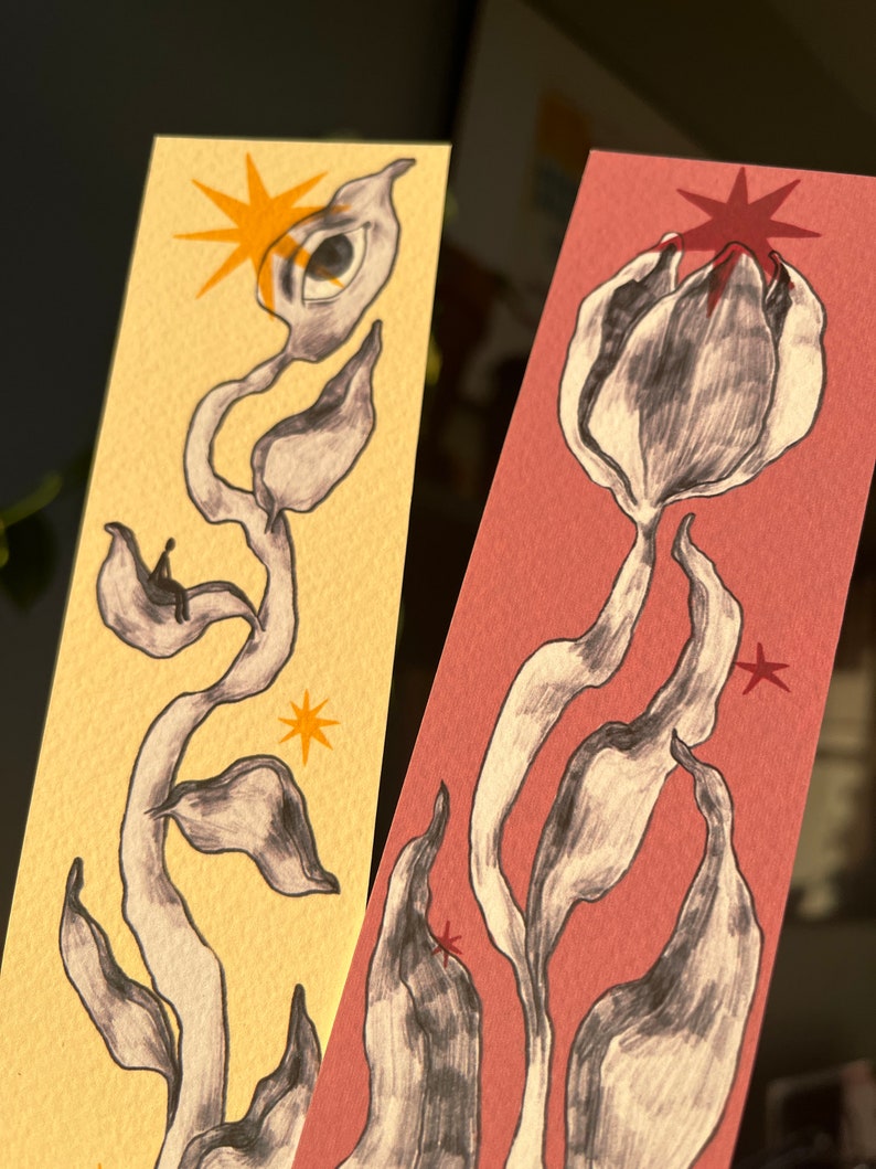 May include: Two hand-painted bookmarks with abstract floral designs. One bookmark is on a yellow background with a black and white flower with an eye and a star. The other bookmark is on a pink background with a black and white flower and a star.