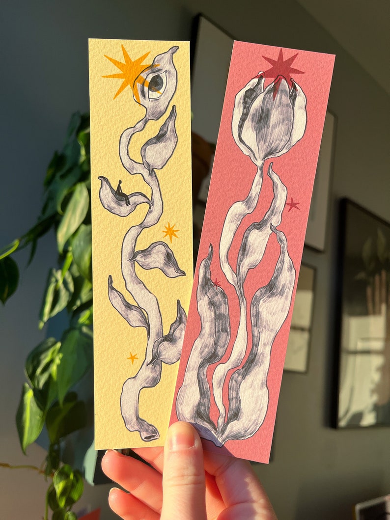 May include: Two hand-painted bookmarks with abstract floral designs. One bookmark has a yellow background with a black and white floral design and a yellow star. The other bookmark has a pink background with a black and white floral design and a red star.