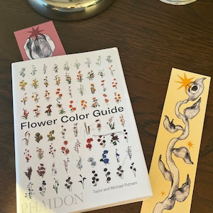 May include: A white book with the title "Flower Color Guide" and the author names "Taylor and Michael Putnam" on the cover. The book is open to a page with illustrations of various flowers in different colors. The book is on a wooden table with two bookmarks.