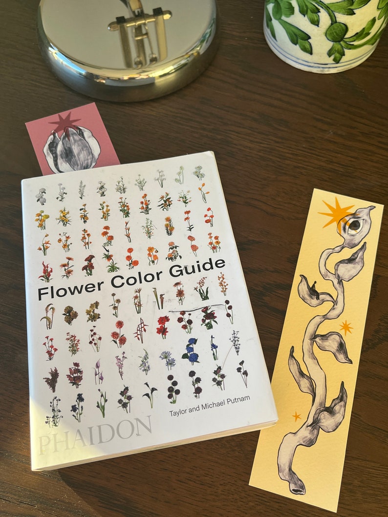 May include: A white book with the title "Flower Color Guide" and the author names "Taylor and Michael Putnam" on the cover. The book is open to a page with illustrations of various flowers in different colors. The book is on a wooden table with two bookmarks.