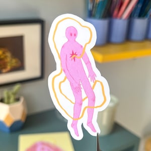 Puede incluir: Una figura rosa con una estrella amarilla en el pecho está rodeada por una línea amarilla. La figura flota en el aire.