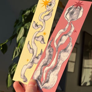 May include: Two bookmarks with hand-drawn illustrations of stylized plants. One bookmark has a yellow background with a black and white plant with a single eye and a yellow star. The other bookmark has a pink background with a black and white plant and a red star.