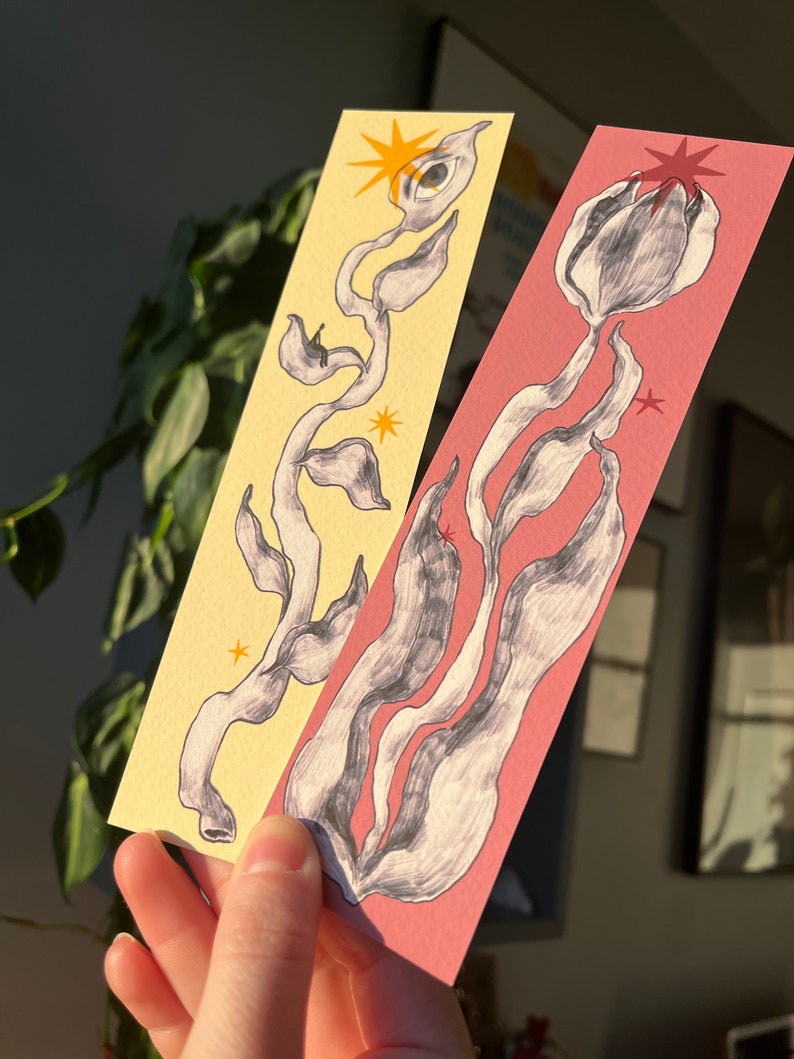 May include: Two bookmarks with hand-drawn illustrations of stylized plants. One bookmark has a yellow background with a black and white plant with a single eye and a yellow star. The other bookmark has a pink background with a black and white plant and a red star.
