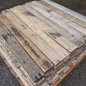 May include: A stack of reclaimed wood planks, weathered and showing signs of age. The planks are arranged in a neat pile, with the grain running in the same direction. The wood is a light brown colour with dark brown streaks and knots.