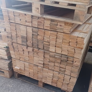 May include: A stack of wooden planks on a pallet. The planks are all the same size and are arranged in a neat, even stack. The planks are light brown in color and have a smooth, finished surface. The pallet is made of wood and has a dark brown color.