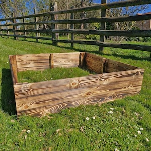 May include: A square wooden raised garden bed with a dark brown, charred finish. The bed is made of thick planks and is sitting on a grassy lawn.