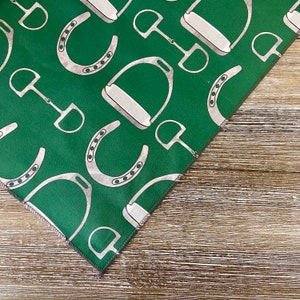 May include: A green fabric with a white pattern of horseshoes and stirrups. The fabric is folded in a triangle shape.