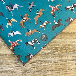 Op de afbeelding: Turkoois stof met een herhalend patroon van paarden en ruiters. De paarden staan in verschillende poses, waaronder springen, rennen en stilstaan. De ruiters dragen kleding in verschillende kleuren.