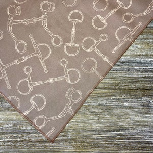 May include: A brown fabric bandana with a white pattern of horse bits. The bandana is folded in a triangle shape and is lying on a wooden surface.