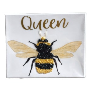 May include: A white canvas with the word "Queen" in gold lettering and a black and gold bee made of beads.