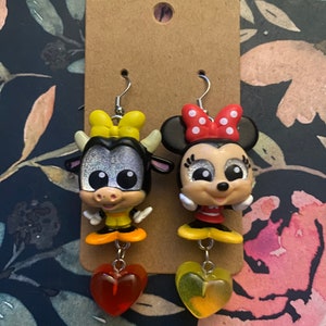 Peut inclure: Une paire de boucles d'oreilles pendantes repr&eacute;sentant une vache et une souris de dessin anim&eacute;. La vache porte un n&oelig;ud jaune et la souris porte un n&oelig;ud rouge &agrave; pois blancs. Les deux personnages ont des yeux brillants et portent des chaussures jaunes. Les boucles d'oreilles sont attach&eacute;es &agrave; des breloques en forme de c&oelig;ur rouge et jaune.