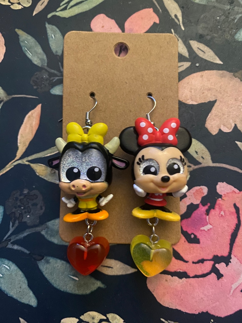 Peut inclure: Une paire de boucles d'oreilles pendantes repr&eacute;sentant une vache et une souris de dessin anim&eacute;. La vache porte un n&oelig;ud jaune et la souris porte un n&oelig;ud rouge &agrave; pois blancs. Les deux personnages ont des yeux brillants et portent des chaussures jaunes. Les boucles d'oreilles sont attach&eacute;es &agrave; des breloques en forme de c&oelig;ur rouge et jaune.