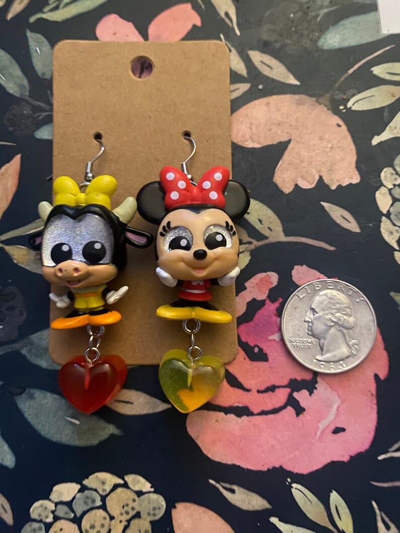 Peut inclure: Une paire de boucles d'oreilles pendantes avec une vache de dessin anim&eacute; et une souris de dessin anim&eacute; avec des pois rouges et blancs. Les boucles d'oreilles ont des breloques en forme de c&oelig;ur jaunes et rouges.