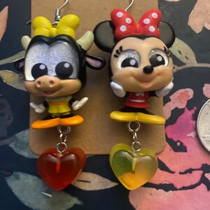 Peut inclure: Une paire de boucles d'oreilles pendantes avec une vache et une souris de dessin anim&eacute;. La vache est noire et blanche avec un n&oelig;ud jaune et la souris porte un n&oelig;ud rouge &agrave; pois blancs. Les deux breloques sont attach&eacute;es &agrave; une cha&icirc;ne argent&eacute;e avec un charme en forme de c&oelig;ur rouge et jaune.