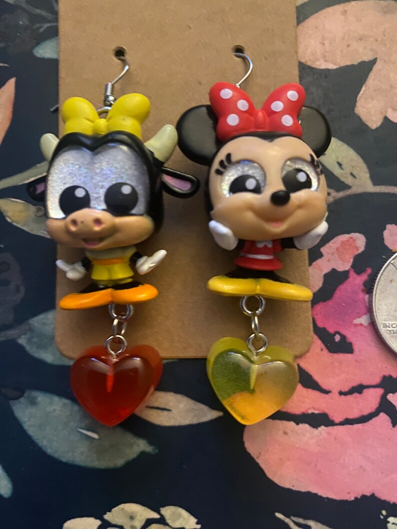 Peut inclure: Une paire de boucles d'oreilles pendantes avec une vache et une souris de dessin anim&eacute;. La vache est noire et blanche avec un n&oelig;ud jaune et la souris porte un n&oelig;ud rouge &agrave; pois blancs. Les deux breloques sont attach&eacute;es &agrave; une cha&icirc;ne argent&eacute;e avec un charme en forme de c&oelig;ur rouge et jaune.