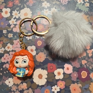 May include: A keychain featuring a cartoon figure with red hair, a blue dress, and a gold key. The keychain has a gold ring and chain, and a fluffy gray pom-pom. The background is a floral pattern.