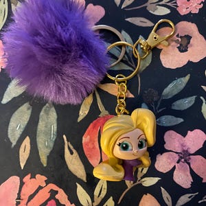 May include: A keychain featuring a cartoon character with blonde hair and a purple dress. The charm is attached to a gold-colored keyring and a purple fluffy pom-pom.