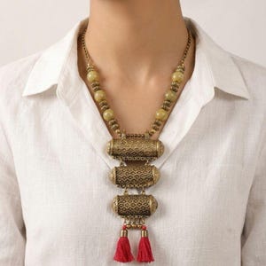 Può includere: Una collana color oro con tre pendenti a forma di barile fantasia e nappe rosse. La collana presenta perline verde oliva e una catena. I pendenti hanno disegni intricati e le nappe aggiungono un tocco di colore al gioiello.