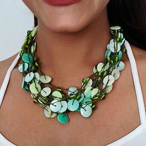 Op de afbeelding: Een meerlagige ketting met groene en witte schijfvormige kralen. De ketting heeft een gelaagd ontwerp met kleine groene kralen. De ketting wordt gedragen over een witte top.