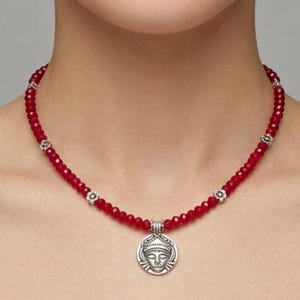 May include: A red beaded necklace with a silver pendant. The pendant features a detailed face design. The necklace has small silver accents and is worn around the neck. The beads are a deep red color.