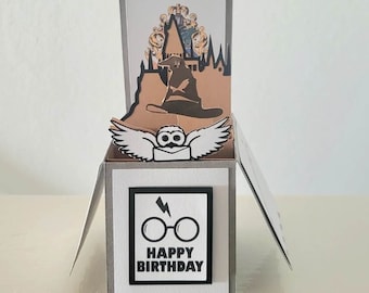 May include: A pop-up card with a Hogwarts castle, a Sorting Hat, and an owl. The card is decorated with a silver border and the words "Happy Birthday" with a lightning bolt and glasses.