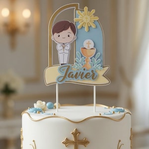 Può includere: Una torta bianca con rifiniture dorate e una croce dorata, decorata con elementi floreali blu e bianchi. La decorazione superiore della torta presenta un ragazzo dei cartoni animati e il nome "Javier". La torta è su un supporto bianco e dorato.