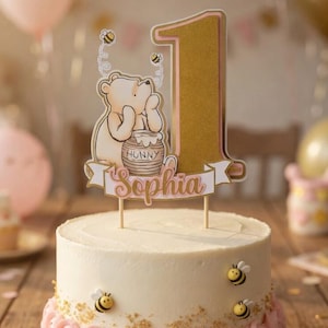 May include: A white iced cake with pink buttercream roses, gold sprinkles, and bee decorations. The cake is topped with a gold number one and a Winnie the Pooh themed topper with the name "Sophia".