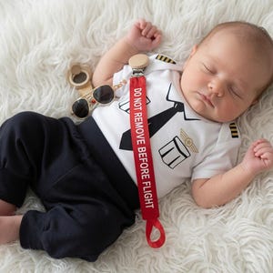 May include: A white short-sleeved bodysuit with a black pilot uniform graphic, black trousers, and a red dummy clip featuring the white text DO NOT REMOVE BEFORE FLIGHT. A pair of small aviator-style sunglasses with dark lenses is included.