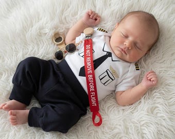 May include: A white short-sleeved bodysuit with a black pilot uniform graphic, black pants, and a red pacifier clip featuring the white text DO NOT REMOVE BEFORE FLIGHT. A pair of small aviator-style sunglasses with dark lenses is included.