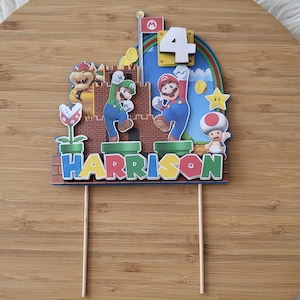 Könnte beinhalten: Ein farbenfroher Tortenaufsatz mit Mario- und Luigi-Figuren, der Zahl 4 und dem Namen "Harrison". Das Design umfasst eine Backsteinmauer, einen Regenbogen und eine Flagge mit einem roten "M"-Symbol. Der Aufsatz ist auf Holzstäben befestigt.