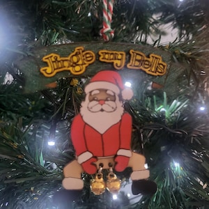 Op de afbeelding: Een houten kerstversiering met de kerstman en de tekst "Jingle my Bells". De versiering is rood, wit en bruin met gouden bellen. Het hangt aan een kerstboom.