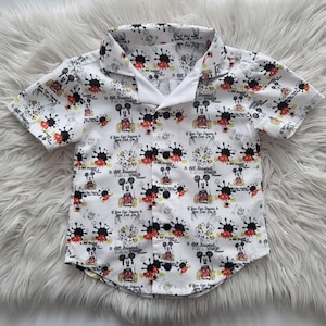 Könnte beinhalten: Weißes, kurzärmeliges Hemd mit Knöpfen und einem schwarzen und weißen Mickey-Mouse-Druck. Das Hemd hat einen weißen Kragen und schwarze Knöpfe. Der Druck zeigt Mickey Mouse in verschiedenen Posen mit dem Text "If You Can Dream It You Can Do It" und "It All Started With A Mouse".