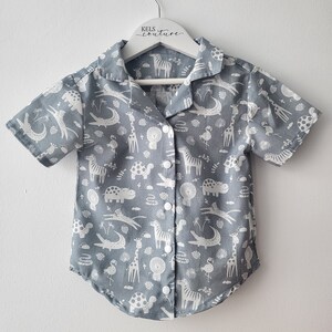 Puede incluir: Una camisa gris de manga corta con botones y un estampado blanco de varios animales, incluyendo jirafas, cebras, hipopótamos y cocodrilos. La camisa tiene cuello y cierre de botones en la parte delantera.