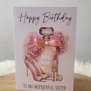 以下が含まれることがあります： 「Happy Birthday」と「To My Wonderful Sister」の文字が入ったバースデーカード。香水瓶、ハイヒール、口紅がピンク色で描かれ、花のアクセントが施されています。
