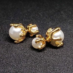 May include: A pair of double-sided earrings featuring two white spherical pearls of different sizes, each encased in a polished gold-tone metal setting with a claw-like design that wraps around the surface of the pearls to secure them in place.