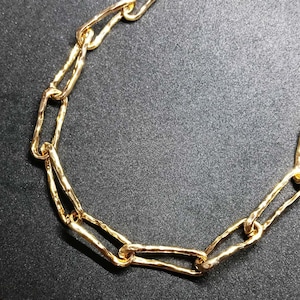May include: A gold-toned chain necklace featuring elongated, rectangular links with a textured, hammered surface. The links are interconnected to form a continuous, flexible chain structure with a polished, reflective finish throughout the piece.