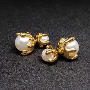 May include: Two pairs of gold-coloured earrings with a pearl design. Each earring features a large, round, white pearl held by a gold-coloured, claw-like setting. The earrings are on a dark, textured surface.