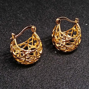 May include: A pair of gold-toned hoop earrings featuring an intricate, openwork lattice design. The earrings have a rounded, basket-like shape with a hinged closure mechanism at the top, showcasing a polished metallic finish throughout the structure.