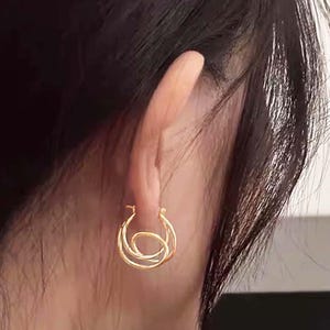 May include: Gold-coloured hoop earrings with an unusual, intertwined design. The earrings are circular, with a smaller, central hoop nested within two larger hoops. The jewellery is shown on a person's ear, with dark hair in the background.