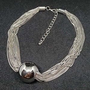 Puede incluir: Un collar plateado de múltiples hilos que presenta numerosas cadenas finas de tipo caja unidas. Una única esfera metálica grande, lisa y reflectante se sitúa como punto focal sobre las cadenas. Incluye un cierre de mosquetón y una cadena extensora.