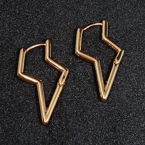 May include: A pair of gold-colored, geometric earrings. Each earring is shaped like a lightning bolt with a hinged closure. The earrings are displayed against a dark, textured background.