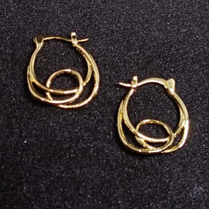 Puede incluir: Un par de pendientes de aro dorados. Cada pendiente presenta un diseño circular con un círculo interior y líneas curvas, creando un efecto en capas. Los pendientes están sobre un fondo oscuro y texturizado.