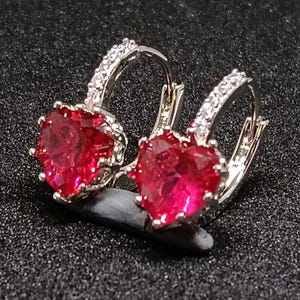 May include: A pair of silver hoop earrings, each with a heart-shaped, bright red gemstone. The hoops are embellished with small, clear gemstones. The earrings are presented on a dark, textured surface.