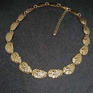 Puede incluir: Un collar de tono dorado que presenta una serie de discos metálicos de formas irregulares interconectados con una superficie martillada y texturizada. La pieza incluye una delicada cadena extensora con una pequeña cuenta redonda en el extremo.