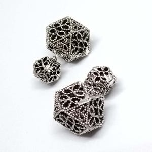 Op de afbeelding: Een paar zilverkleurige oorbellen met een dubbelzijdig ontwerp met twee geometrische, kubusvormige onderdelen. Elk onderdeel is vervaardigd met een ingewikkeld, opengewerkt filigraanpatroon dat een delicaat, kantachtig uiterlijk creëert.