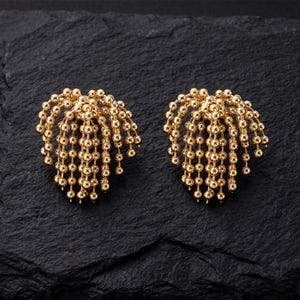 May include: A pair of gold-toned stud earrings featuring a cluster of cascading ball chains. Each earring is composed of multiple strands of small, reflective gold spheres that fan out from a central point, creating a textured, fringe-like appearance.