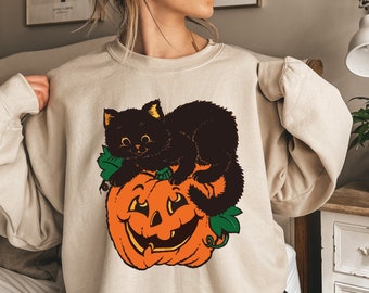 May include: A beige sweatshirt with a black cat sitting on an orange pumpkin with green leaves. The cat is looking to the right and has a white face.